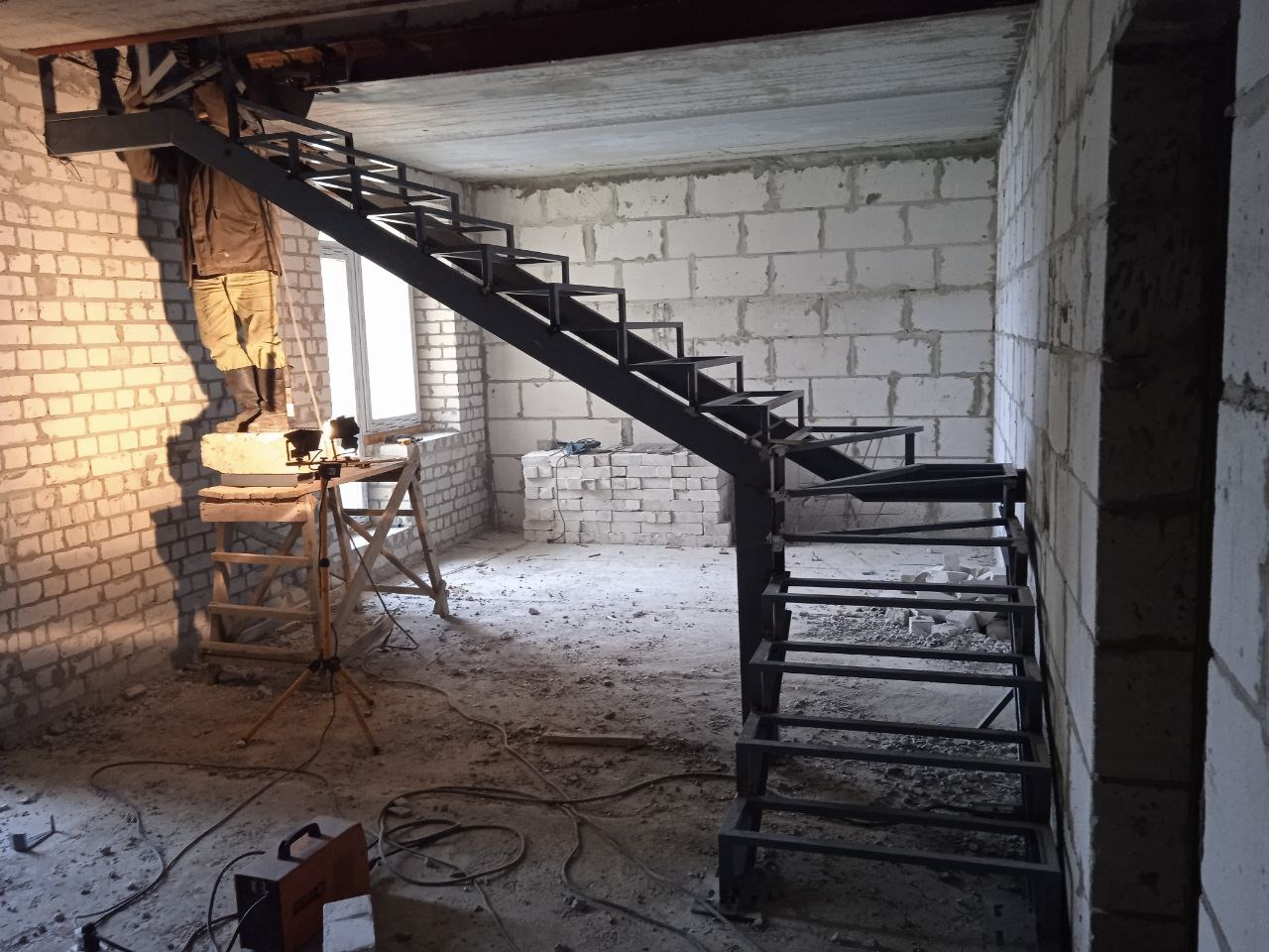 Montaż metalowych schodów w surowym wnętrzu. Widoczny spawacz na podeście, ceglane ściany i sprzęt spawalniczy. Schody o konstrukcji stalowej, jeszcze bez wykończenia.