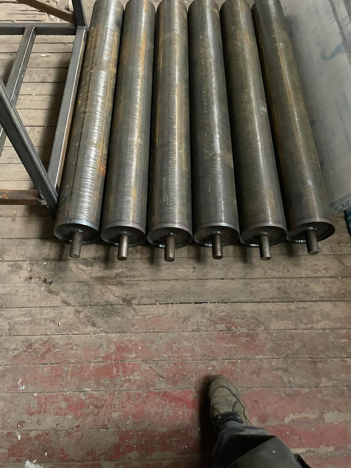Sześć metalowych, cylindrycznych rolek na podłodze z desek, widoczne ślady spawania, fragment konstrukcji stalowej w tle. Surowy, warsztatowy klimat.