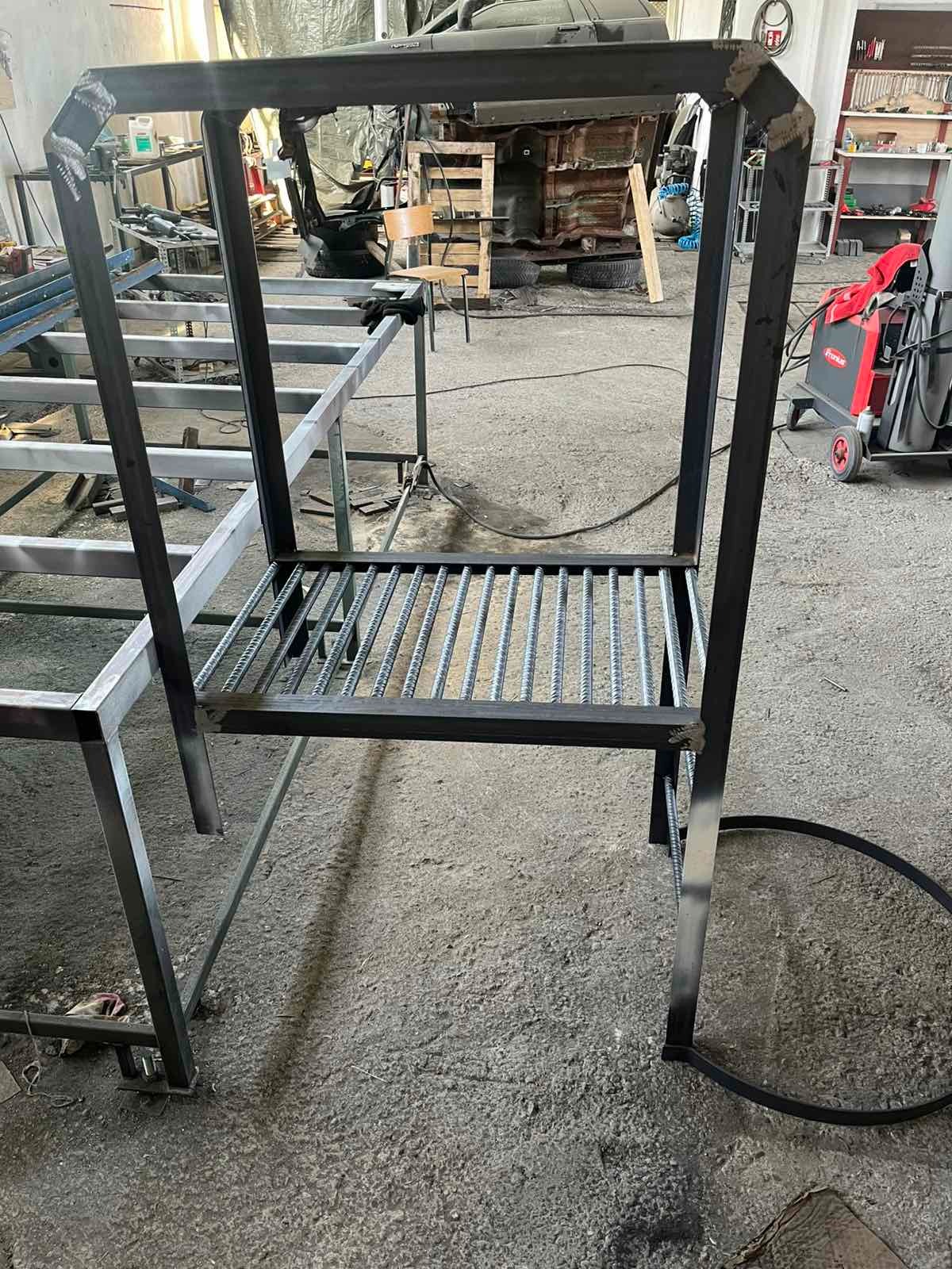 Konstrukcja metalowa z prętami, prawdopodobnie w trakcie tworzenia, widoczna spawarka i warsztatowe otoczenie. Surowy, industrialny charakter.