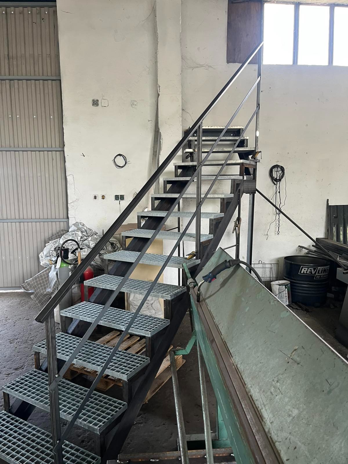 Metalowe schody kratowe z poręczą w surowym wnętrzu warsztatu, widoczne narzędzia ślusarskie i materiały. Schody prowadzą do okna na górze.