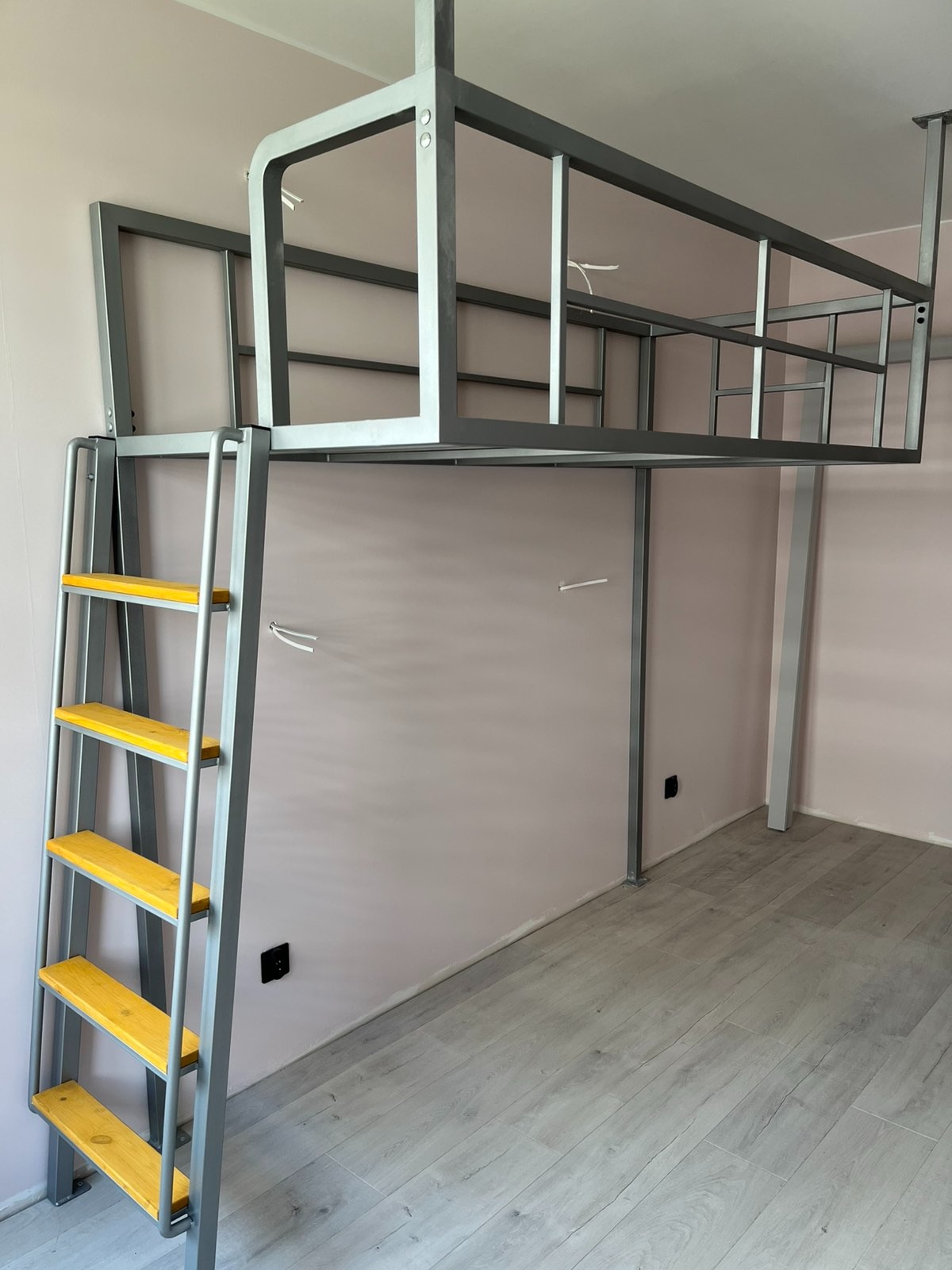 Metalowa konstrukcja łóżka piętrowego z drabinką o żółtych szczeblach, zawieszona pod sufitem, w pokoju z panelami podłogowymi i ścianami w odcieniach różu.