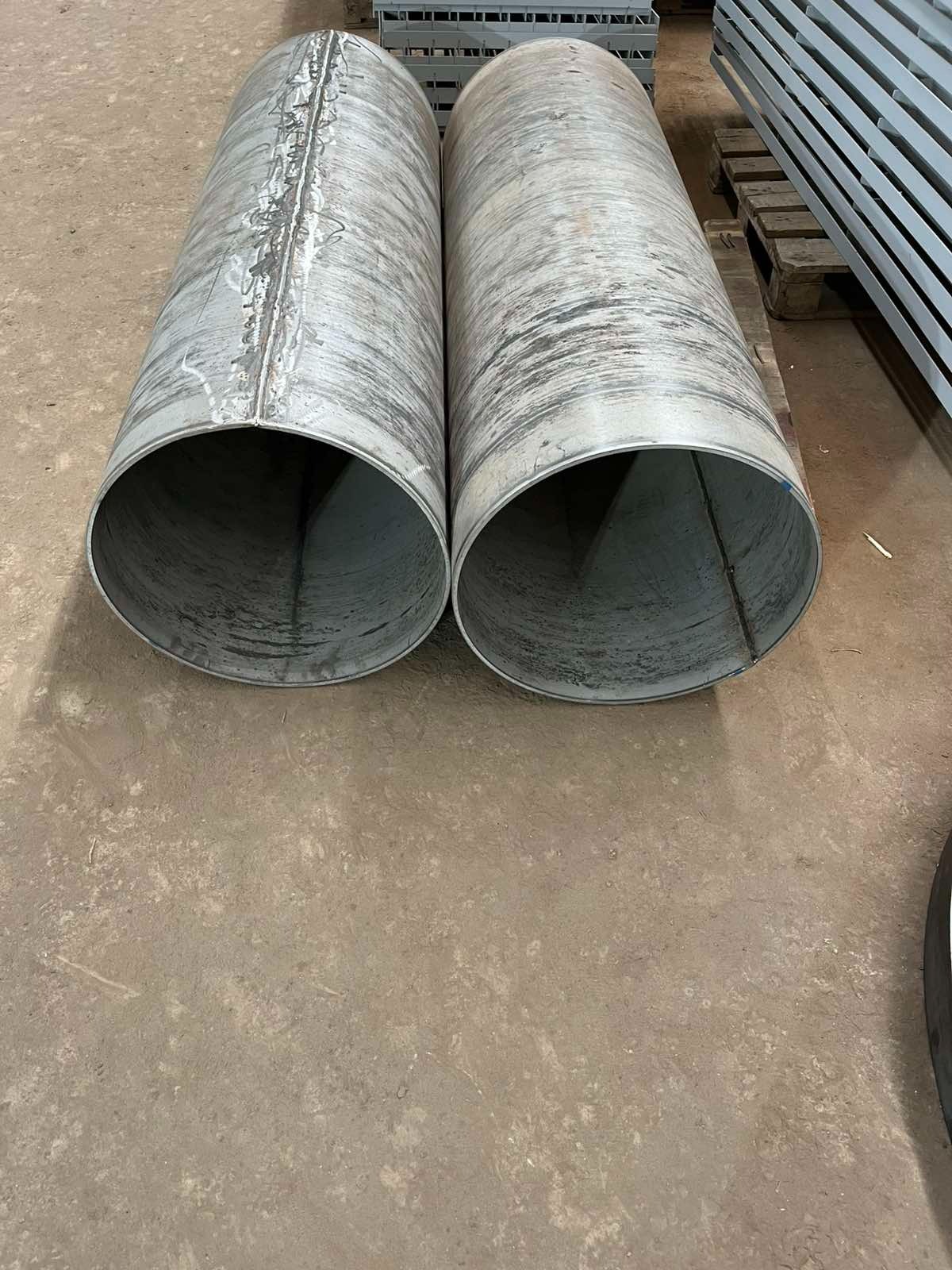 Dwie spawane rury ze stali nierdzewnej leżące na podłodze warsztatu, widoczne ślady po spawaniu, w tle elementy konstrukcji stalowej na palecie.