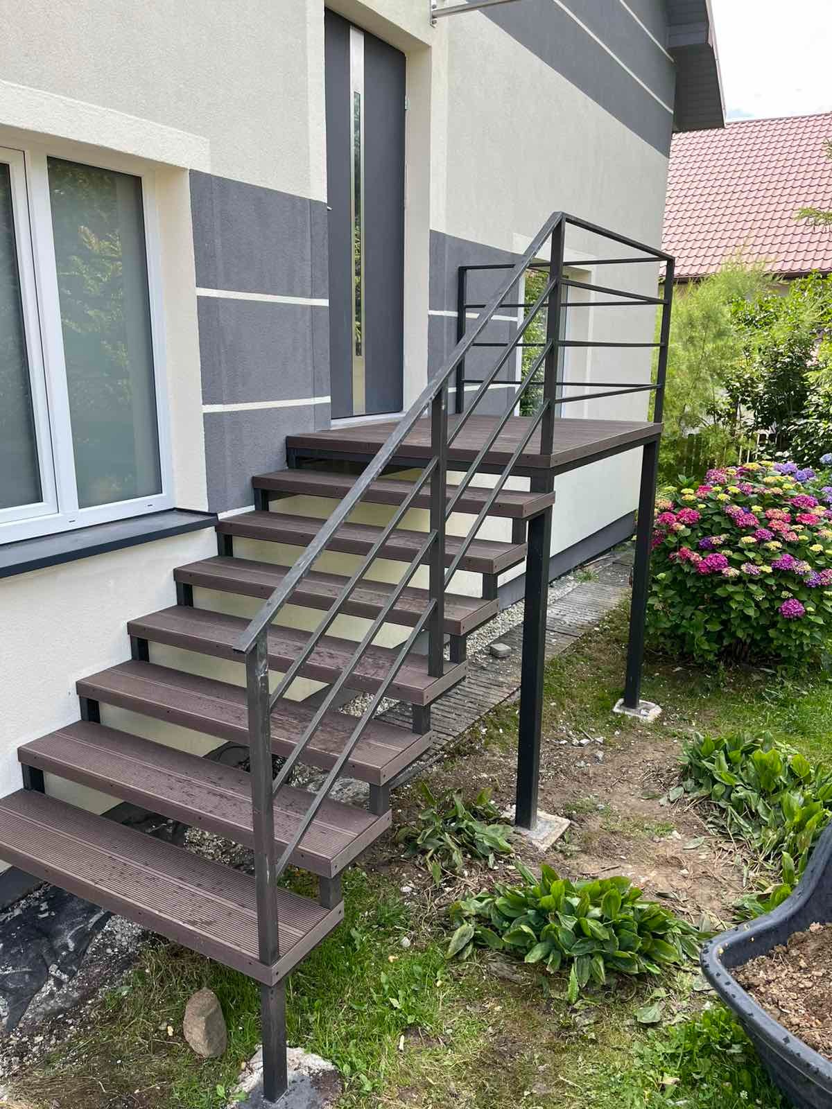 Zewnętrzne schody z metalową poręczą prowadzące do drzwi domu. Stopnie z desek kompozytowych. Widok z dołu, z ogrodem w tle i fragmentem elewacji budynku.