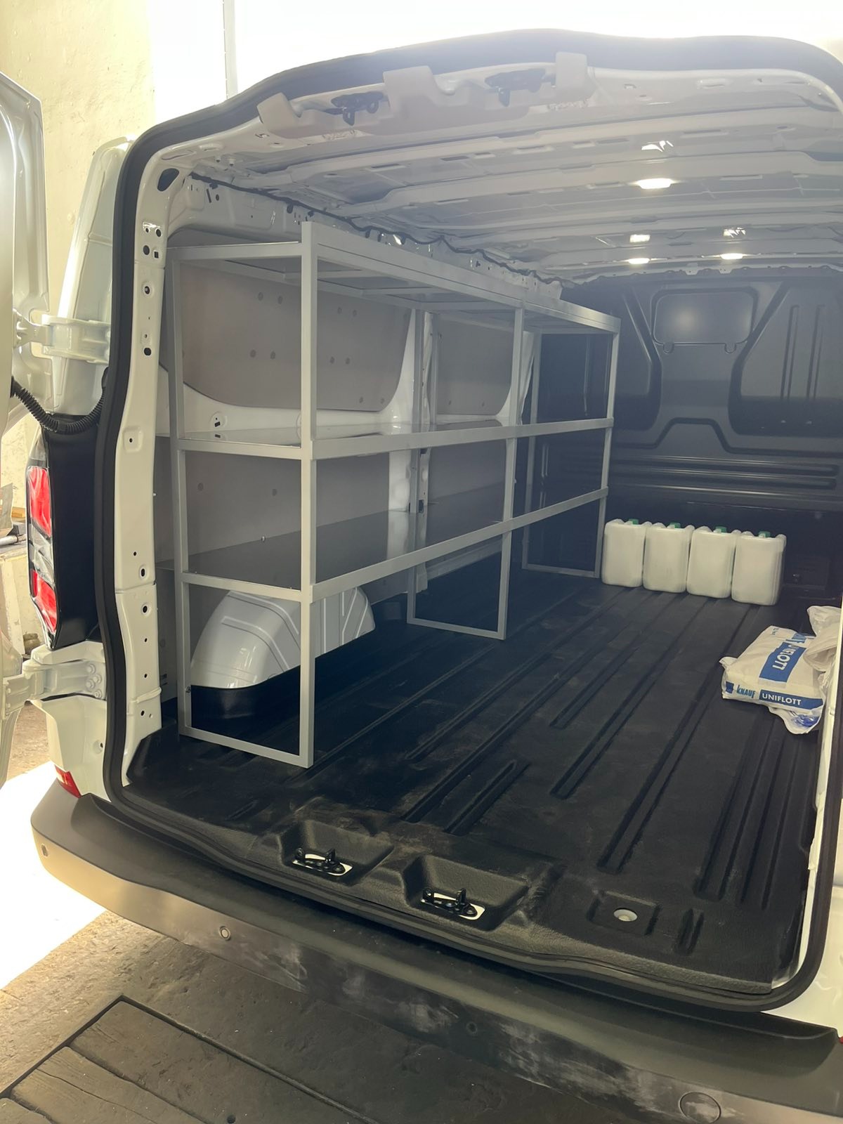 Wnętrze furgonetki z zamontowanym, metalowym regałem na narzędzia i materiały budowlane. Widoczne białe kanistry i worek z zaprawą.