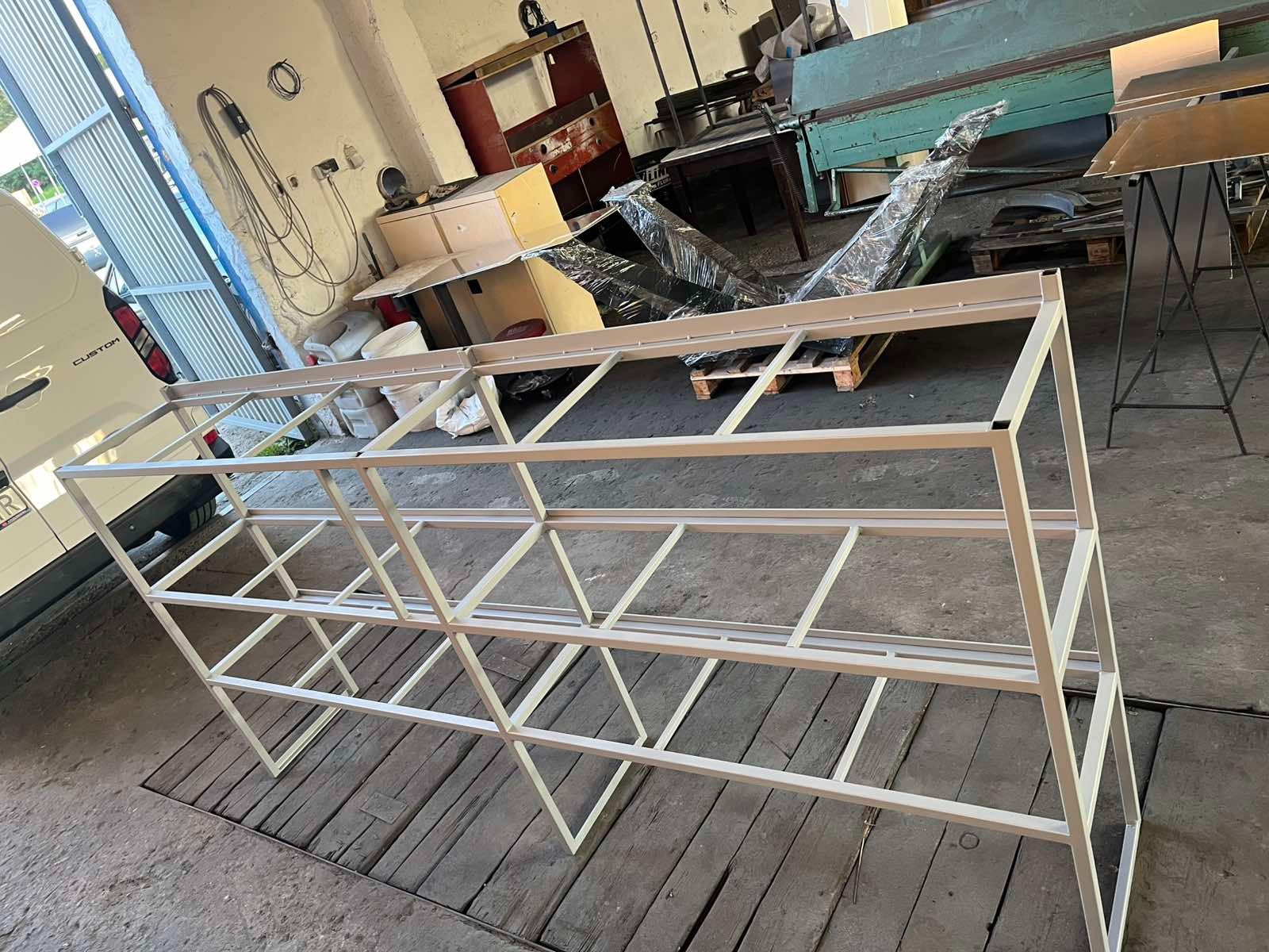 Biała, metalowa konstrukcja regału z kwadratowych profili, spawana, widoczna w warsztacie na tle maszyny do obróbki blachy i częściowo widocznego białego busa.