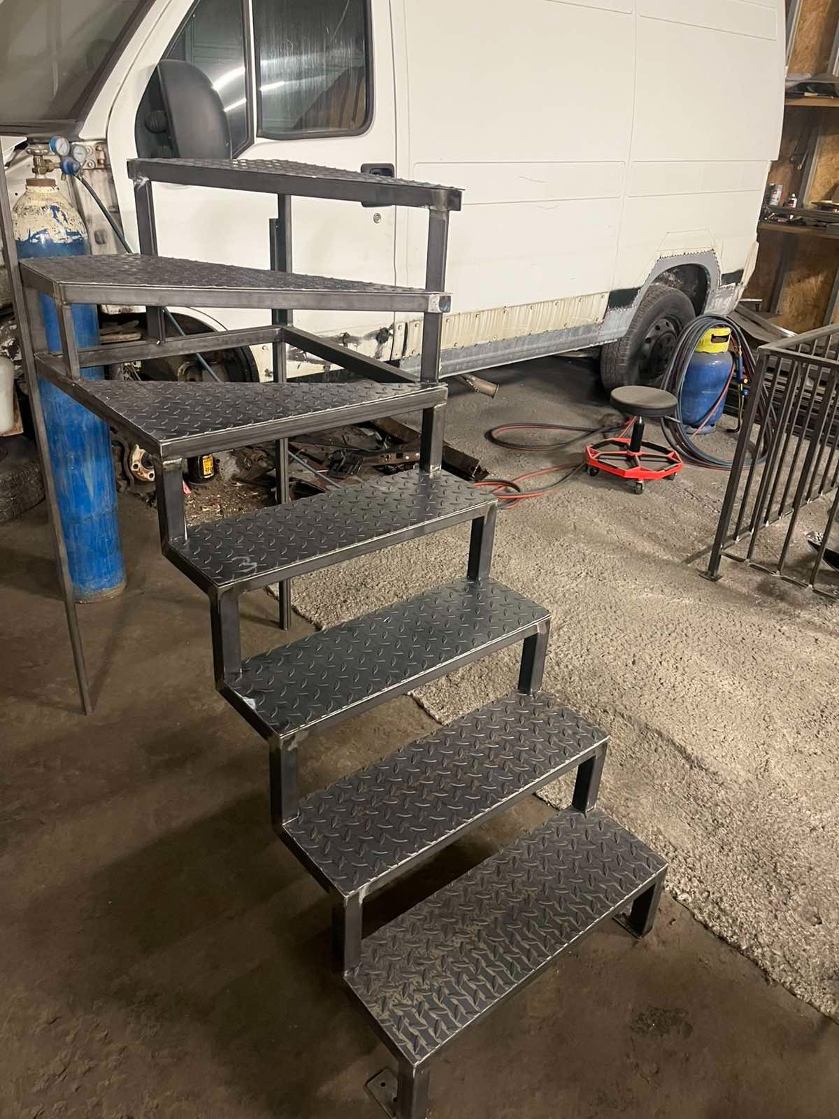 Surowe, metalowe schody z blachy ryflowanej w warsztacie, z widocznym spawem i elementami wyposażenia, na tle białego busa.