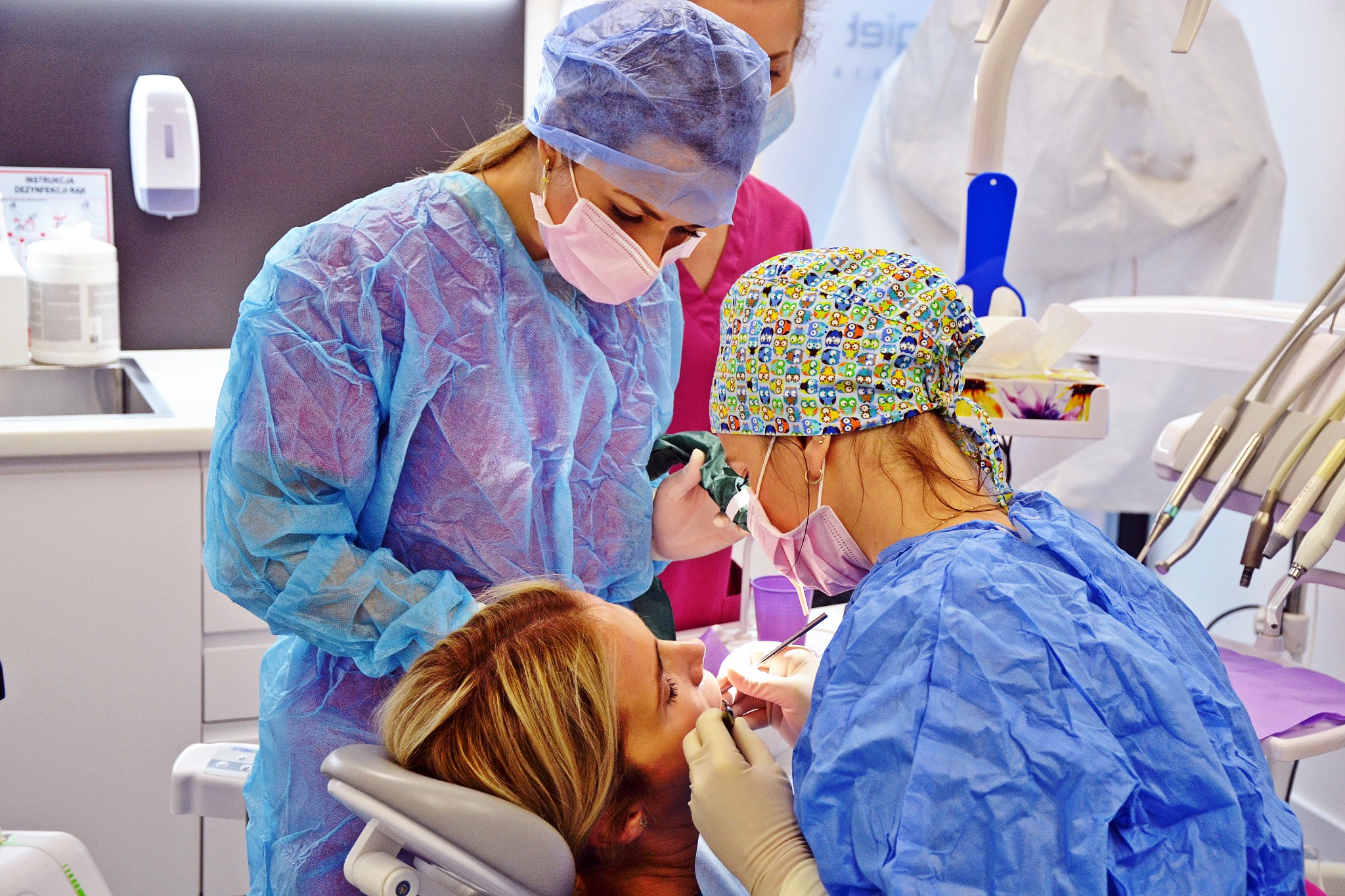 Zespół stomatologiczny w trakcie zabiegu u pacjentki. Lekarze w strojach ochronnych, oświetlenie dentystyczne i narzędzia. Widoczna blond kobieta na fotelu dentystycznym.