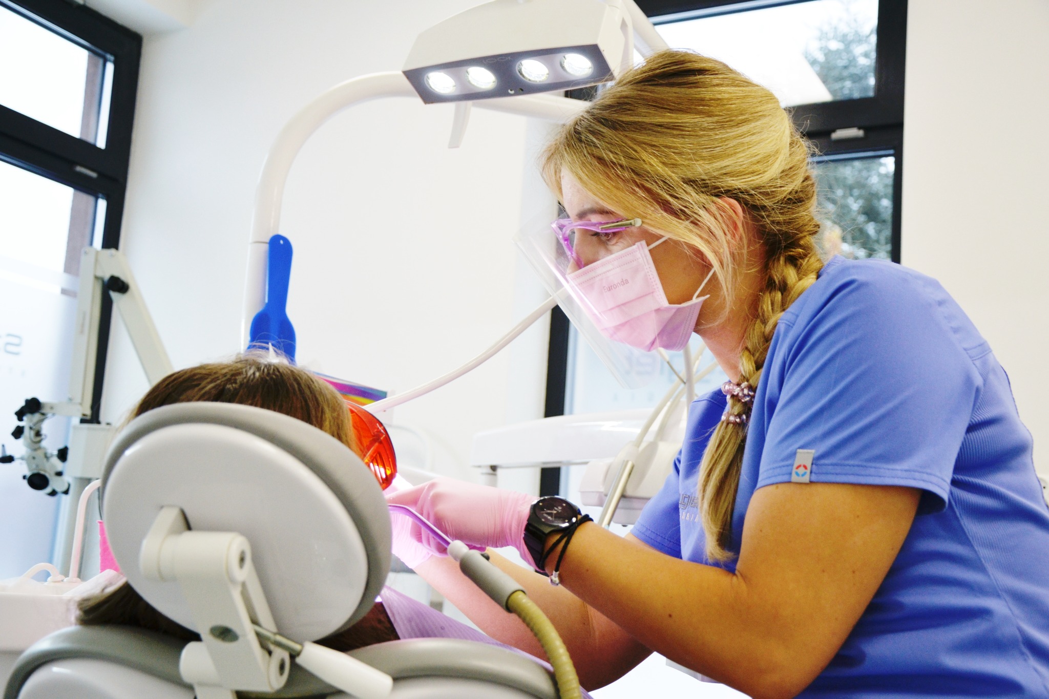 Dentystka w różowej masce i okularach ochronnych, pochylona nad pacjentem, używa narzędzi w jasnym gabinecie. Widoczny reflektor i sprzęt stomatologiczny.