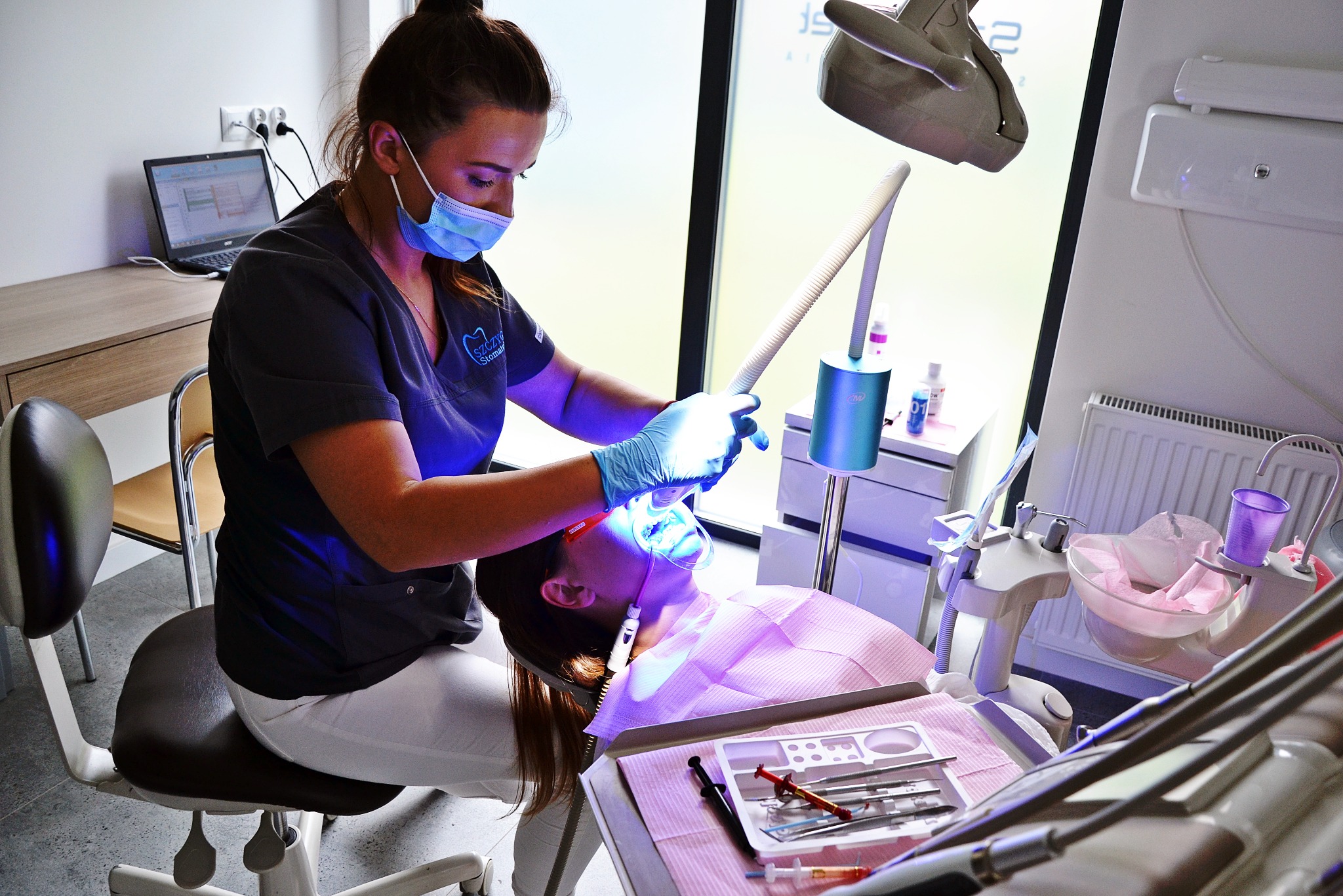 Dentystka w trakcie zabiegu, oświetlenie stomatologiczne, narzędzia na tacy. Widoczny laptop z harmonogramem wizyt w tle.