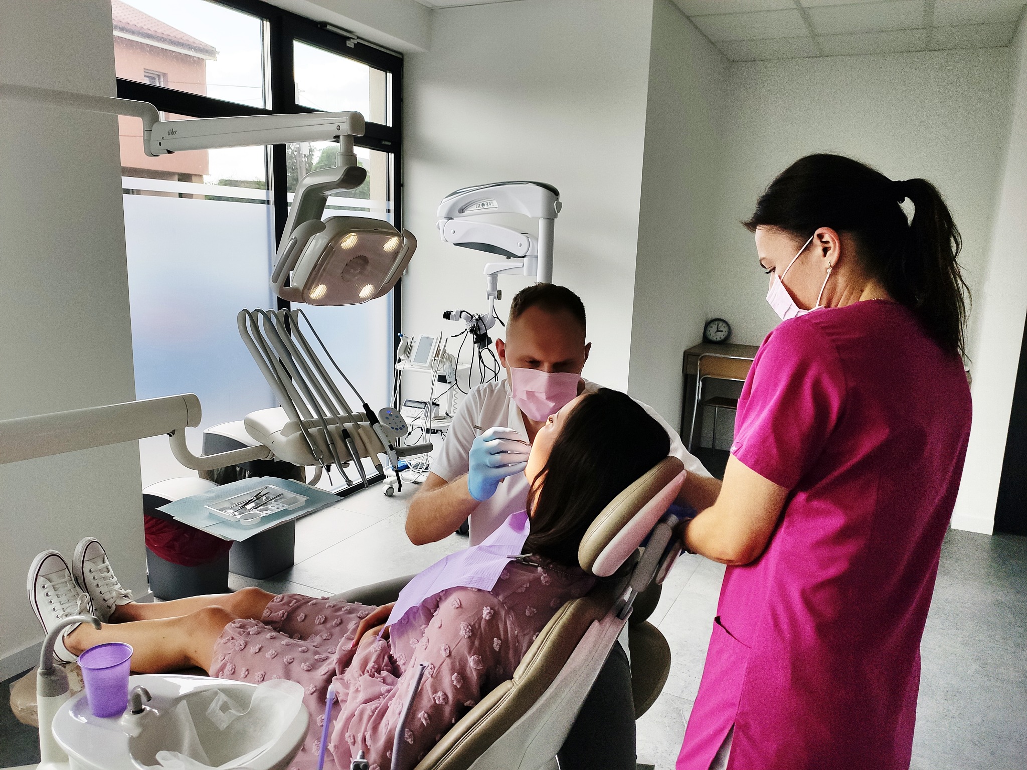 Pacjentka w różowej sukience na fotelu dentystycznym podczas zabiegu. Lekarz i asystentka w maseczkach. Nowoczesny gabinet z dużym oknem i specjalistycznym sprzętem.
