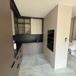DM Invest Damian Zaremba - Nowoczesna kuchnia w trakcie montażu: szare szafki dolne i górne z czarnymi akcentami, wbudowany piekarnik, marmurowa podłoga, widoczne przewody i narzędzia.