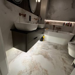 DM Invest Damian Zaremba - Nowoczesna łazienka z dwoma umywalkami nablatowymi, ciemną szafką pod umywalką z szufladami, owalnymi lustrami z podświetleniem, wanną wolnostojącą i marmurową podłogą.