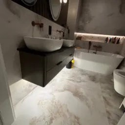 Nowoczesna łazienka z dwoma umywalkami nablatowymi, ciemną szafką pod umywalką z szufladami, owalnymi lustrami z podświetleniem, wanną wolnostojącą i marmurową podłogą.