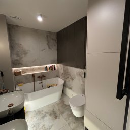 DM Invest Damian Zaremba - Nowoczesna łazienka z owalną wanną, umywalkami nablatowymi i zabudową meblową w odcieniach bieli i szarości, wykończona płytkami imitującymi marmur.