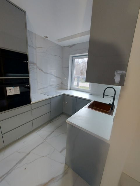 Nowoczesna kuchnia z szarymi frontami, białym blatem i marmurową ścianą oraz podłogą, z czarną armaturą i miedzianym zlewem.