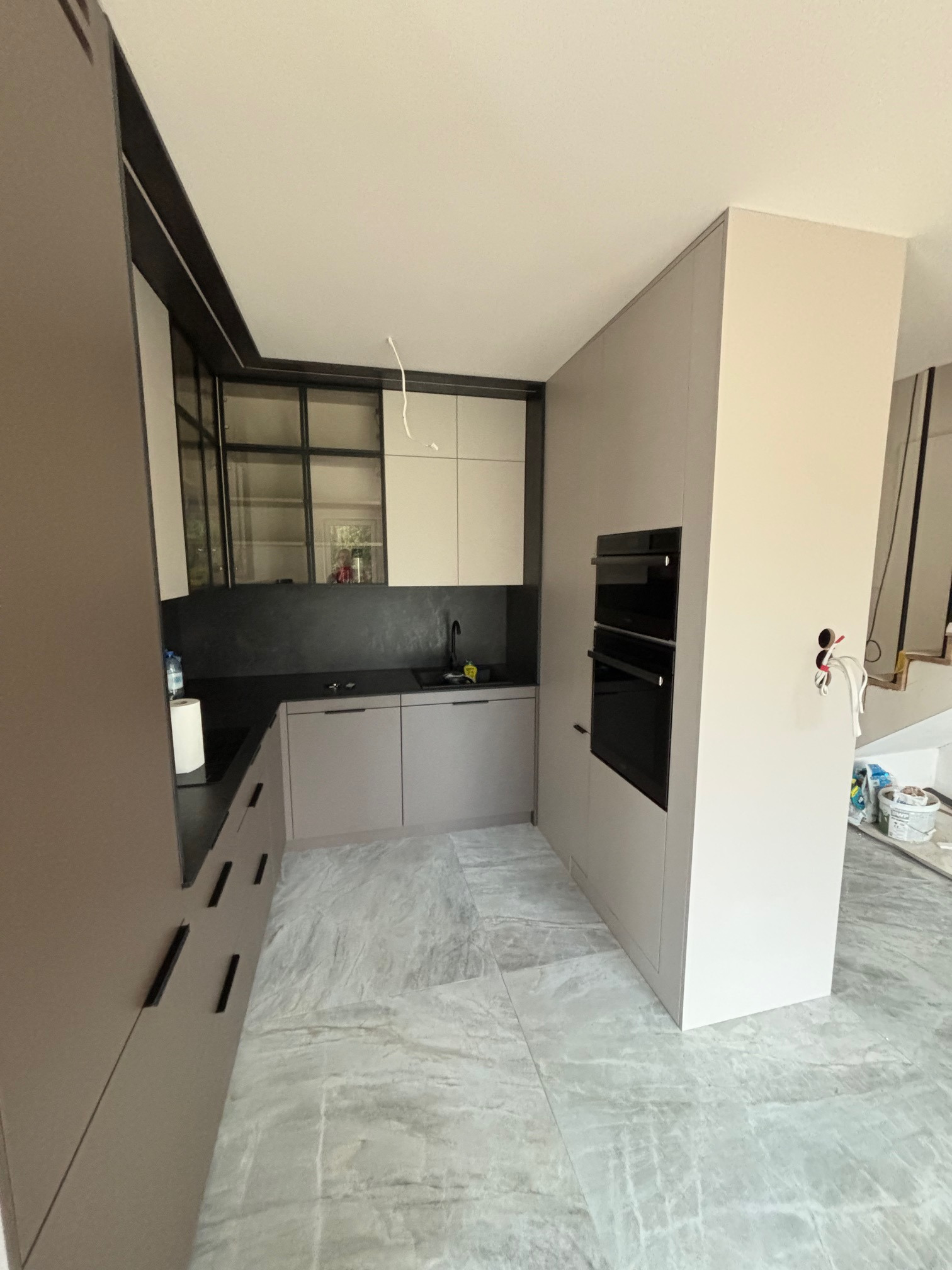 Nowoczesna kuchnia w trakcie montażu: szare szafki dolne i górne z czarnymi akcentami, wbudowany piekarnik, marmurowa podłoga, widoczne przewody i narzędzia.