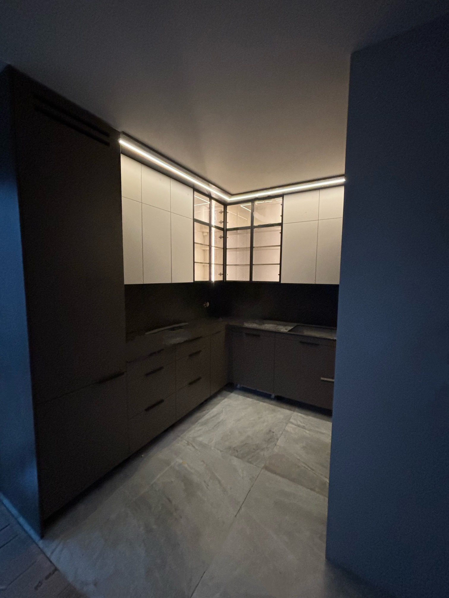 Nowoczesna kuchnia z szarymi frontami szafek dolnych i białymi górnymi szafkami z czarnymi ramkami, oświetlenie LED pod szafkami górnymi, szara podłoga z płytek.