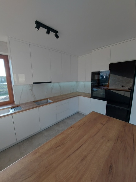 Nowoczesna kuchnia z białymi szafkami, drewnianym blatem i wyspą, oświetleniem punktowym na suficie i marmurowym panelem ściennym.