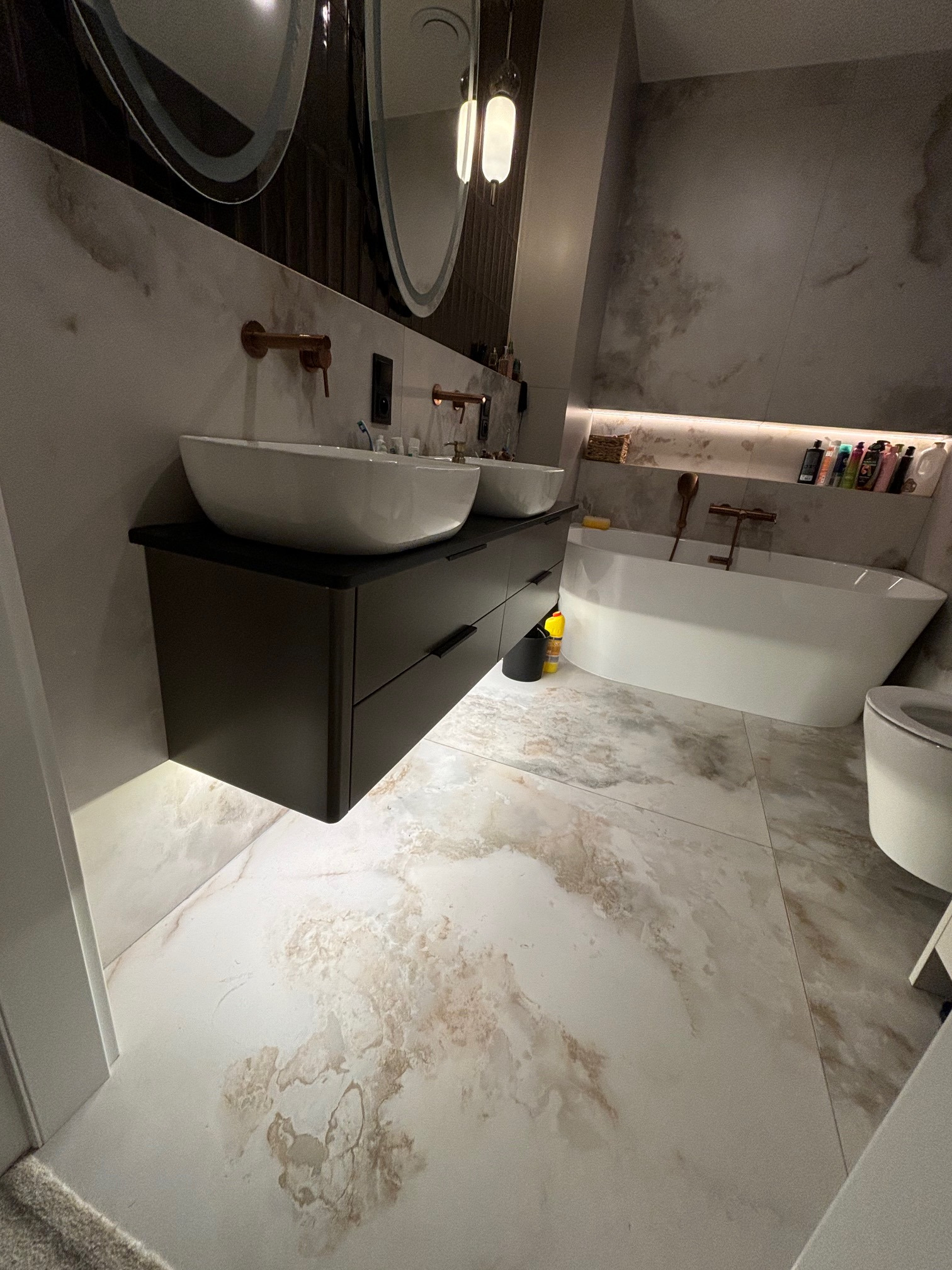 Nowoczesna łazienka z dwoma umywalkami nablatowymi, ciemną szafką pod umywalką z szufladami, owalnymi lustrami z podświetleniem, wanną wolnostojącą i marmurową podłogą.