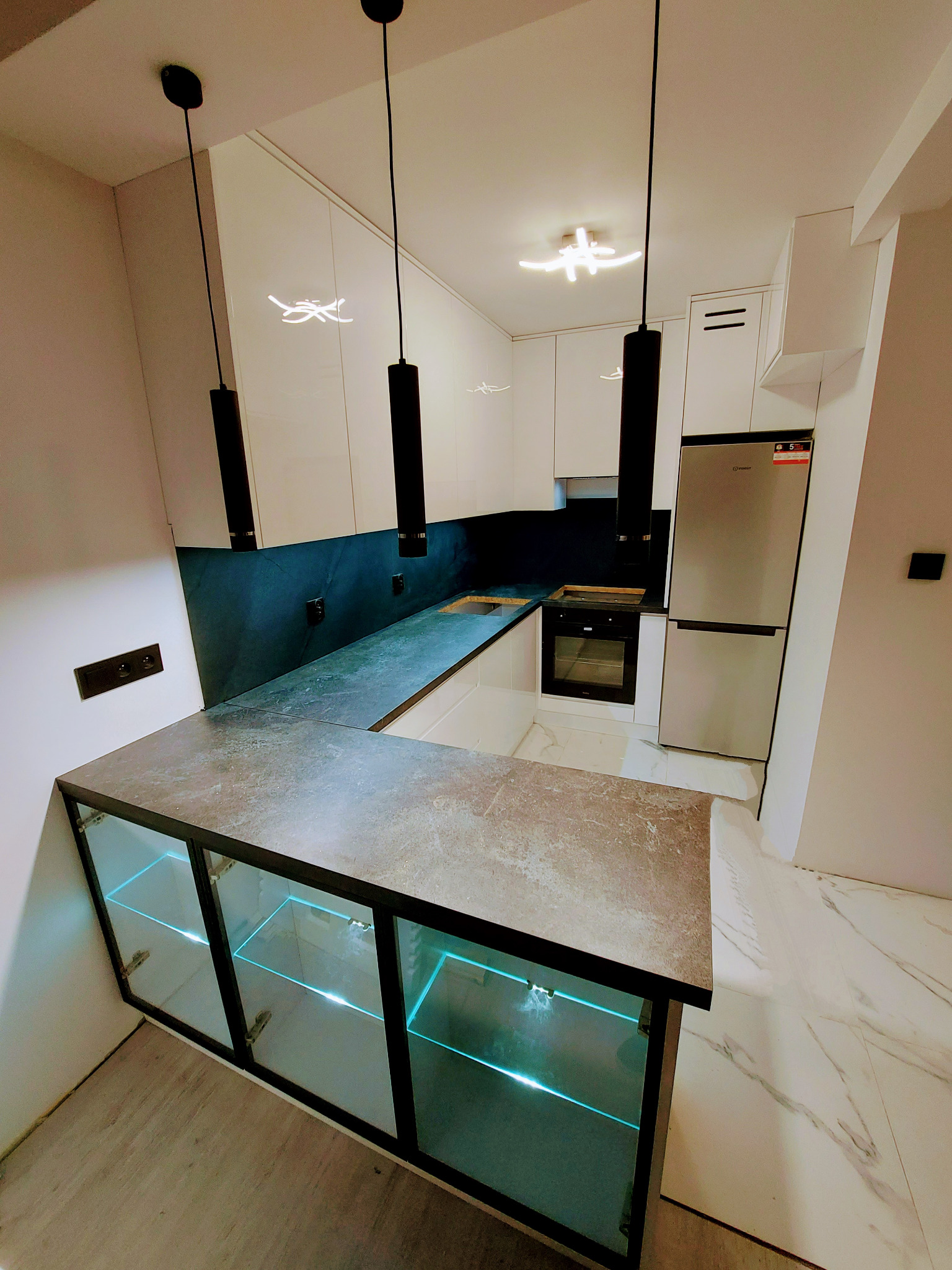 Nowoczesna kuchnia z białymi szafkami, ciemnym blatem i podświetlanymi szklanymi frontami barku, oświetlona wiszącymi lampami i sufitowym plafonem.