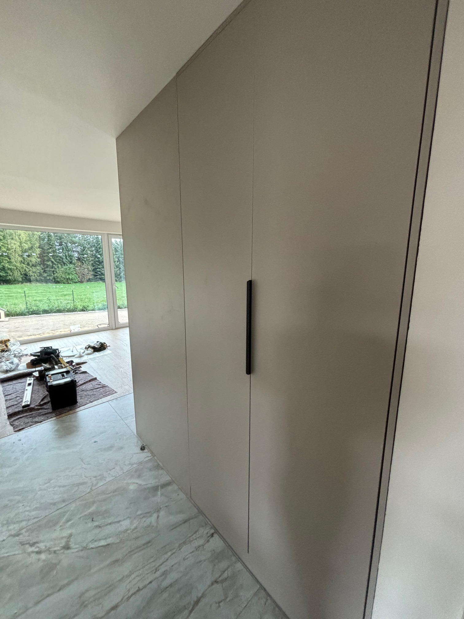 Szara szafa wnękowa z czarnymi, podłużnymi uchwytami, widok z boku podczas montażu, narzędzia na podłodze, duże okno z widokiem na zieleń w tle.