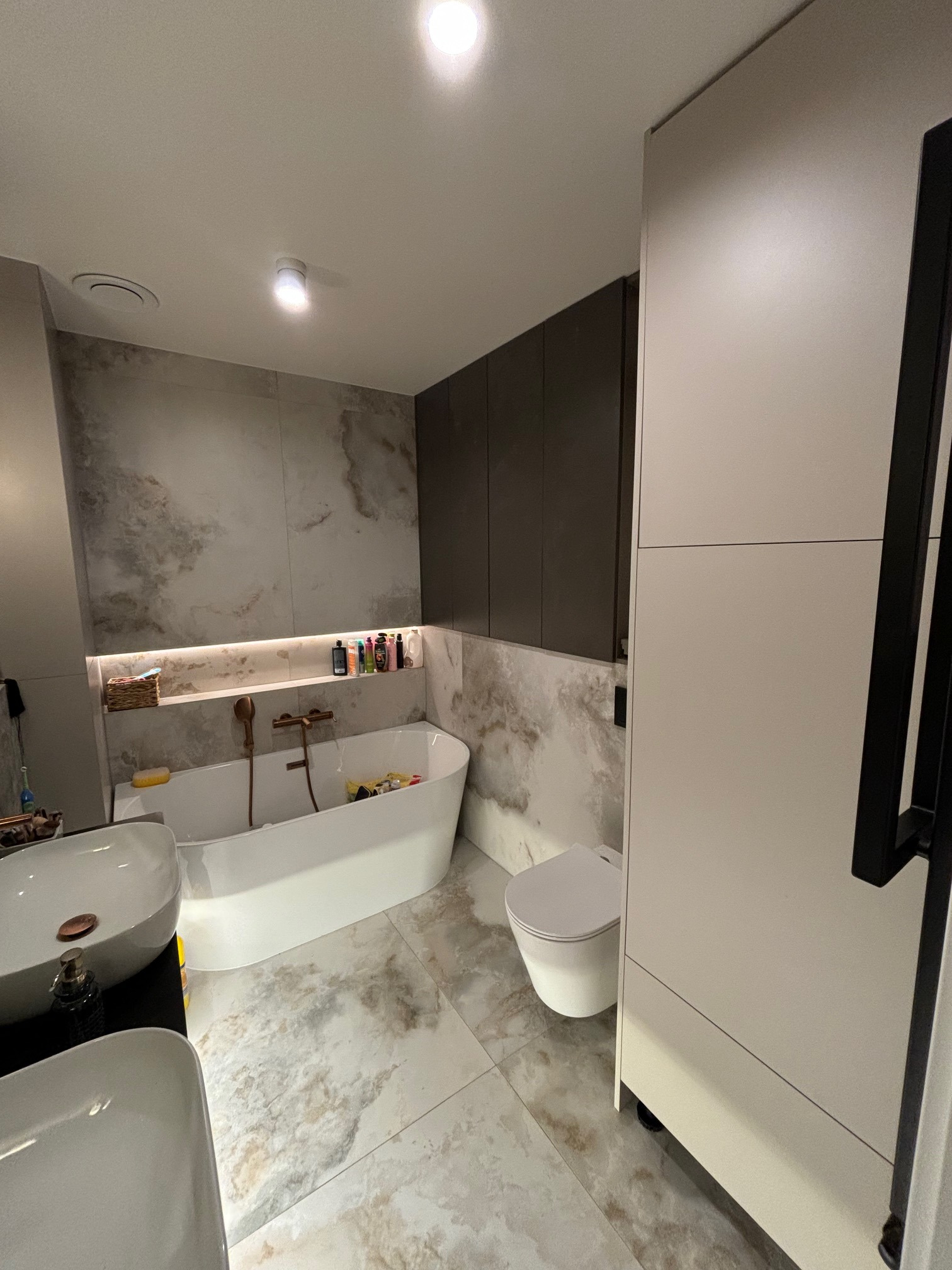 Nowoczesna łazienka z owalną wanną, umywalkami nablatowymi i zabudową meblową w odcieniach bieli i szarości, wykończona płytkami imitującymi marmur.