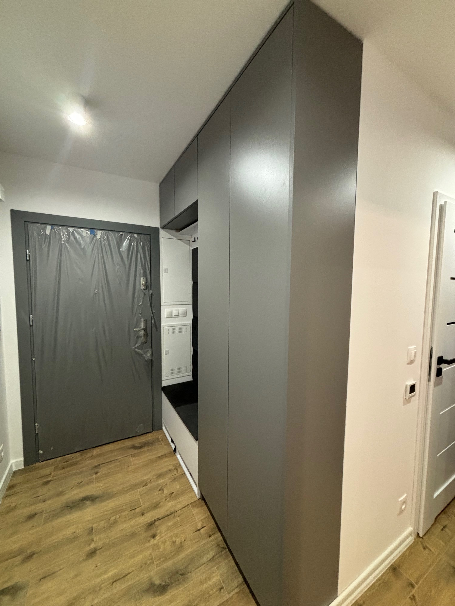 Szara zabudowa meblowa w przedpokoju z siedziskiem i szafkami, drzwi wejściowe zabezpieczone folią ochronną, podłoga imitująca drewno.