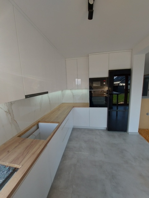 Nowoczesna biała kuchnia z drewnianym blatem i czarną lodówką, widok na fragment blatu przygotowanego pod montaż zlewu.