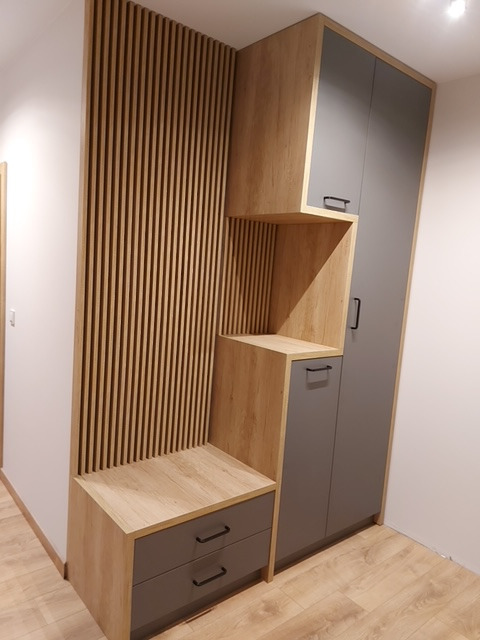 Nowoczesna zabudowa przedpokoju w Radomsku: szafa z szarymi frontami, siedzisko z szufladami i dekoracyjny panel z drewnianych listewek, całość w odcieniach drewna i szarości.