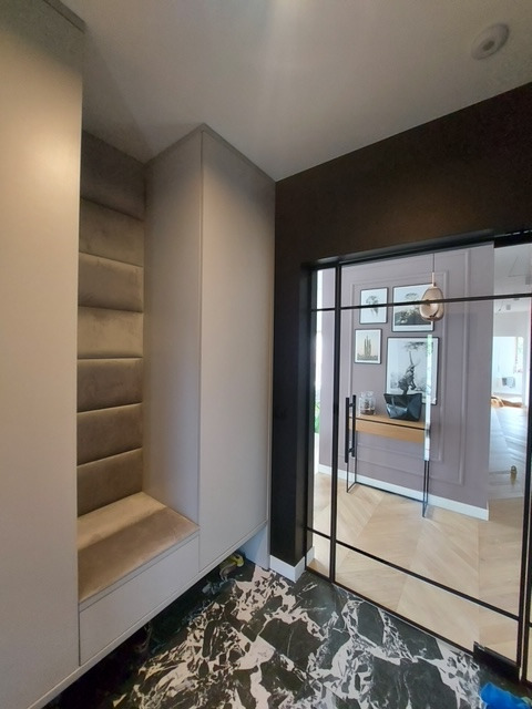 Wnętrze przedpokoju z zabudowaną szafą wnękową z siedziskiem obitym welurem, marmurową posadzką i szklanymi drzwiami przesuwnymi z czarnymi szprosami.