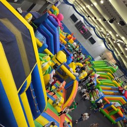 DAGMARA BARWIŃSKA-TRZECIAK RATOWNIK MEDYCZNY - Wielopoziomowy, kolorowy park dmuchany z licznymi przeszkodami i bawiącymi się dziećmi, widoczny z góry, oświetlony jasnym światłem hali.