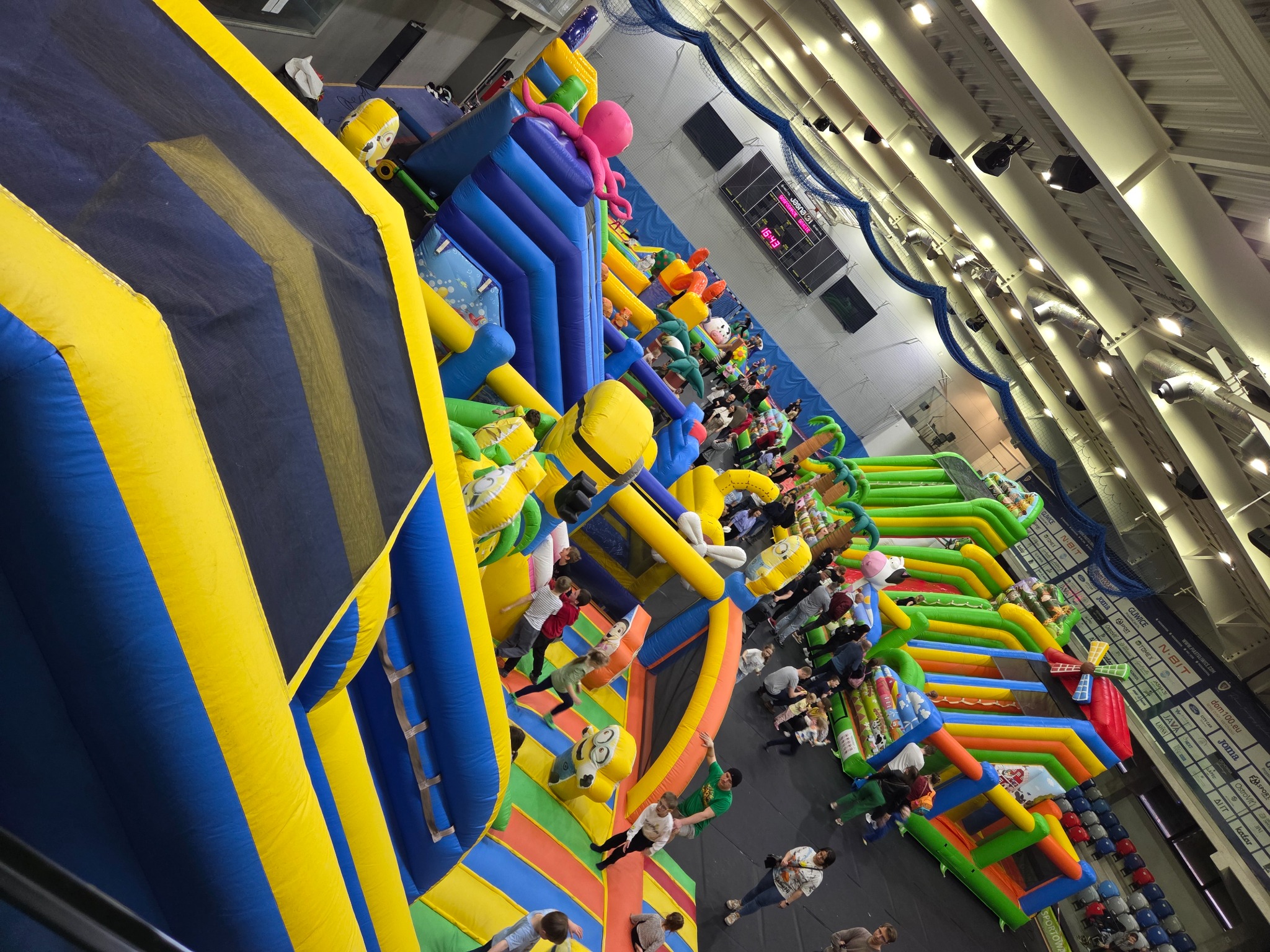 Wielopoziomowy, kolorowy park dmuchany z licznymi przeszkodami i bawiącymi się dziećmi, widoczny z góry, oświetlony jasnym światłem hali.