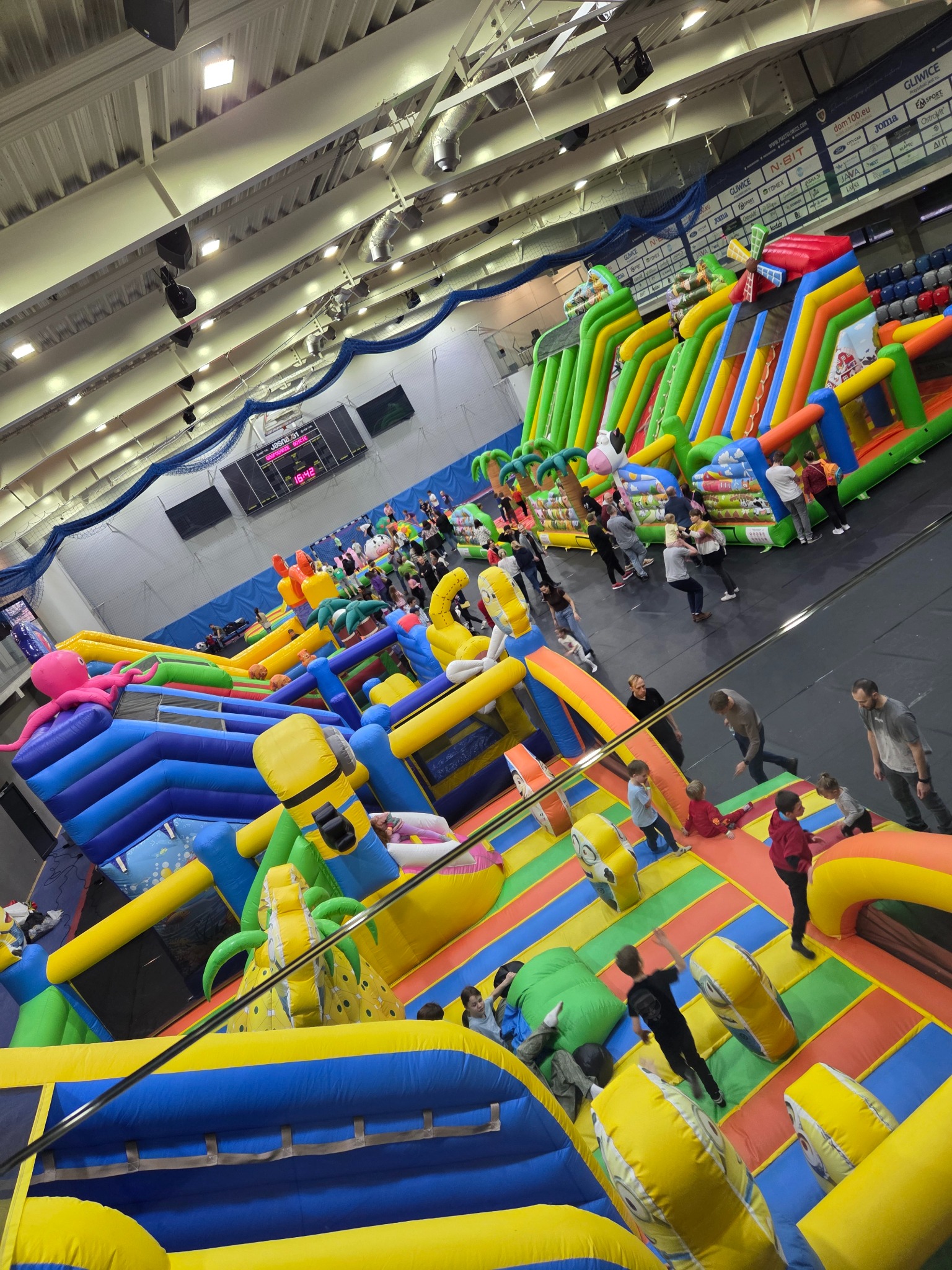 Wielki, kolorowy park rozrywki z dmuchanych atrakcji w hali sportowej, z widocznymi dziećmi i dorosłymi korzystającymi z konstrukcji.