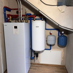 Eco Synergia - Biały, nowoczesny piec grzewczy marki Nibe z podłączonymi rurami w kolorach niebieskim i czerwonym, zbiornik buforowy oraz naczynia przeponowe w pomieszczeniu z wykończonymi płytkami podłogowymi...