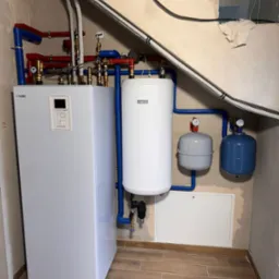 Biały, nowoczesny piec grzewczy marki Nibe z podłączonymi rurami w kolorach niebieskim i czerwonym, zbiornik buforowy oraz naczynia przeponowe w pomieszczeniu z wykończonymi płytkami podłogowymi...