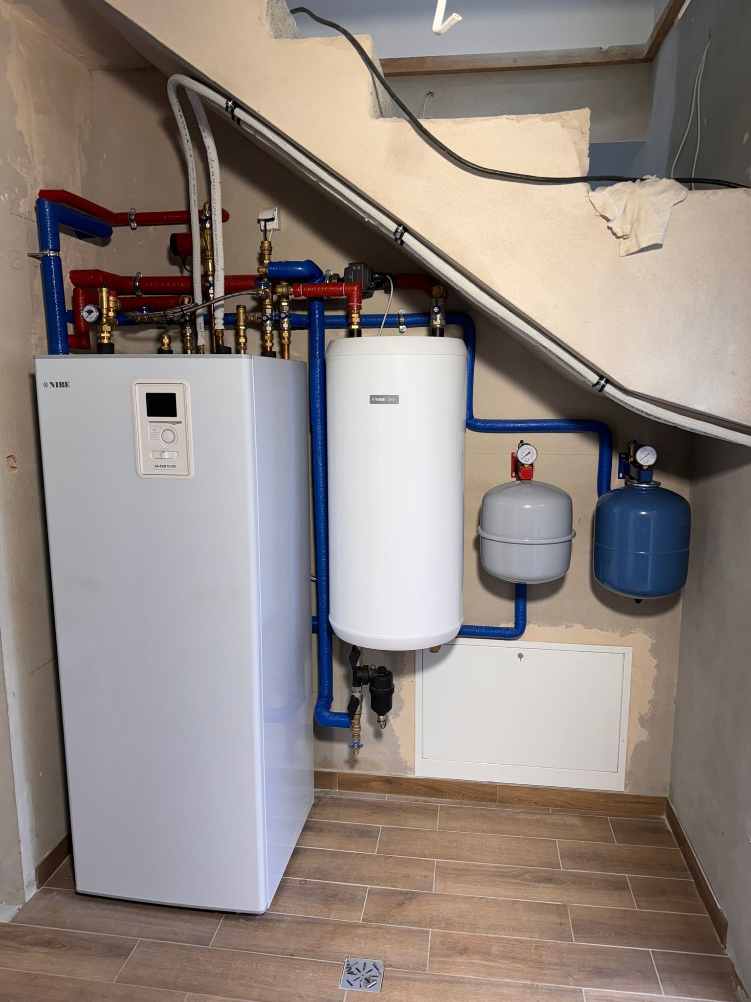 Biały, nowoczesny piec grzewczy marki Nibe z podłączonymi rurami w kolorach niebieskim i czerwonym, zbiornik buforowy oraz naczynia przeponowe w pomieszczeniu z wykończonymi płytkami podłogowymi...