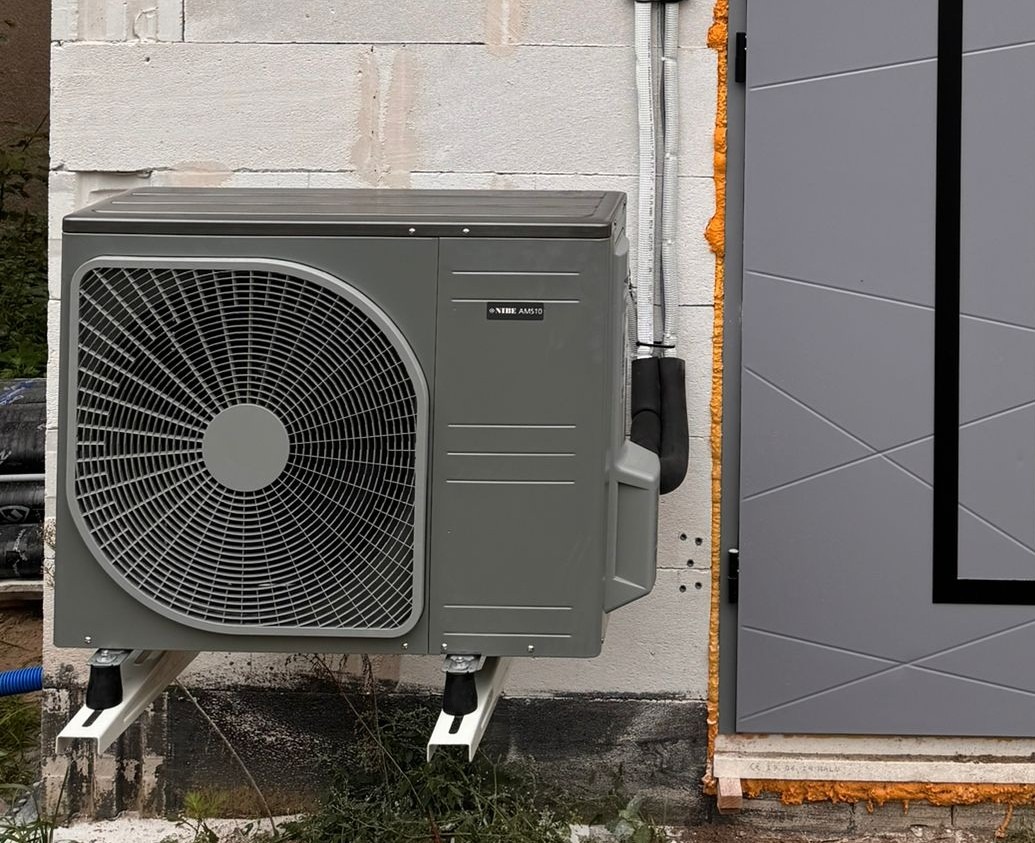 Szara pompa ciepła Nibe AMS10 zamontowana na białej ścianie z betonu komórkowego, widoczne rury instalacyjne i fragment szarego okna z pianką montażową.