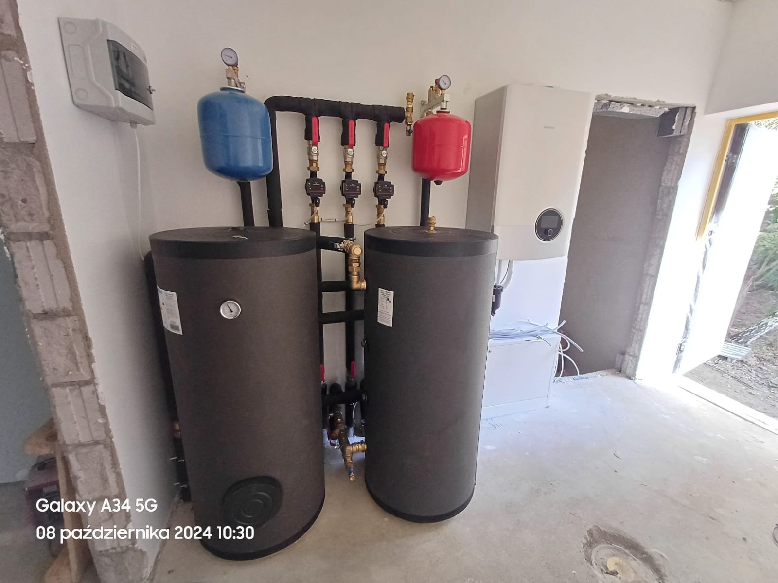 Dwa pionowe zbiorniki buforowe z niebieskim i czerwonym naczyniem wzbiorczym, podłączone do kolektora z zaworami, obok wiszący piec gazowy w surowym pomieszczeniu.