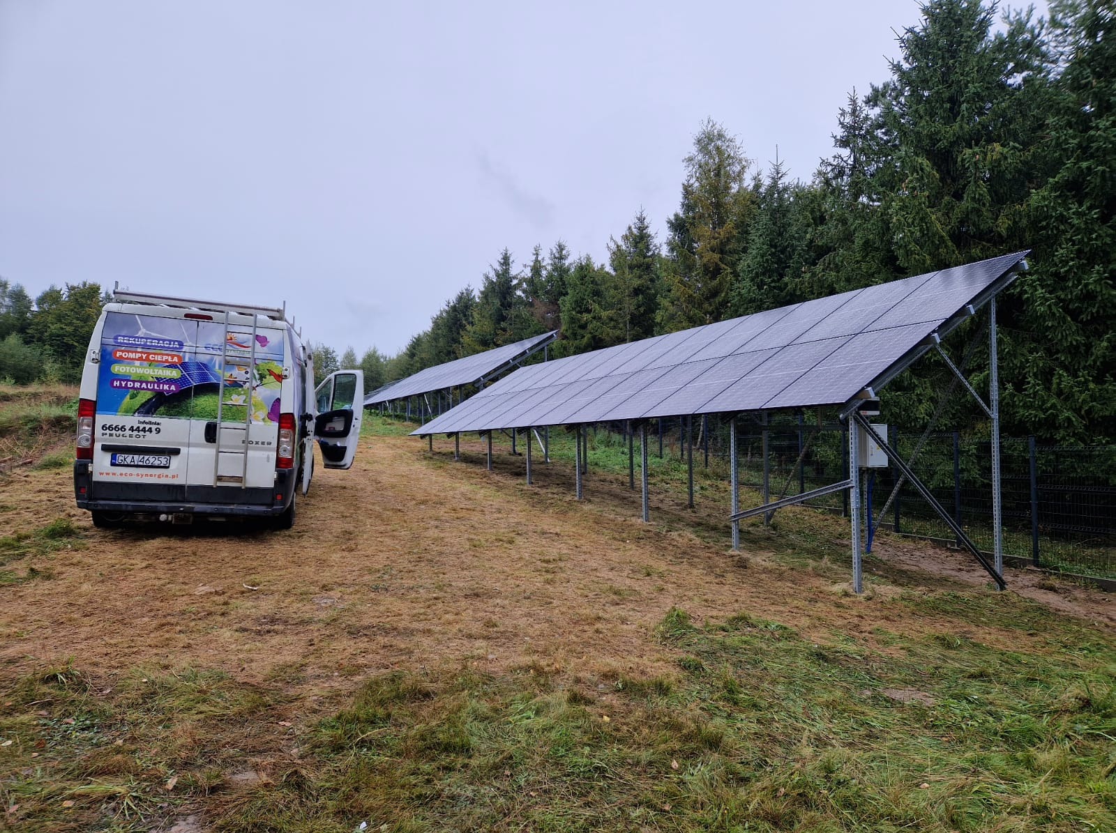 Dwie rzędy wolnostojących paneli słonecznych na konstrukcji gruntowej, widoczne z perspektywy zza białego samochodu dostawczego z otwartymi drzwiami, zaparkowanego na trawiastym terenie obok lasu.