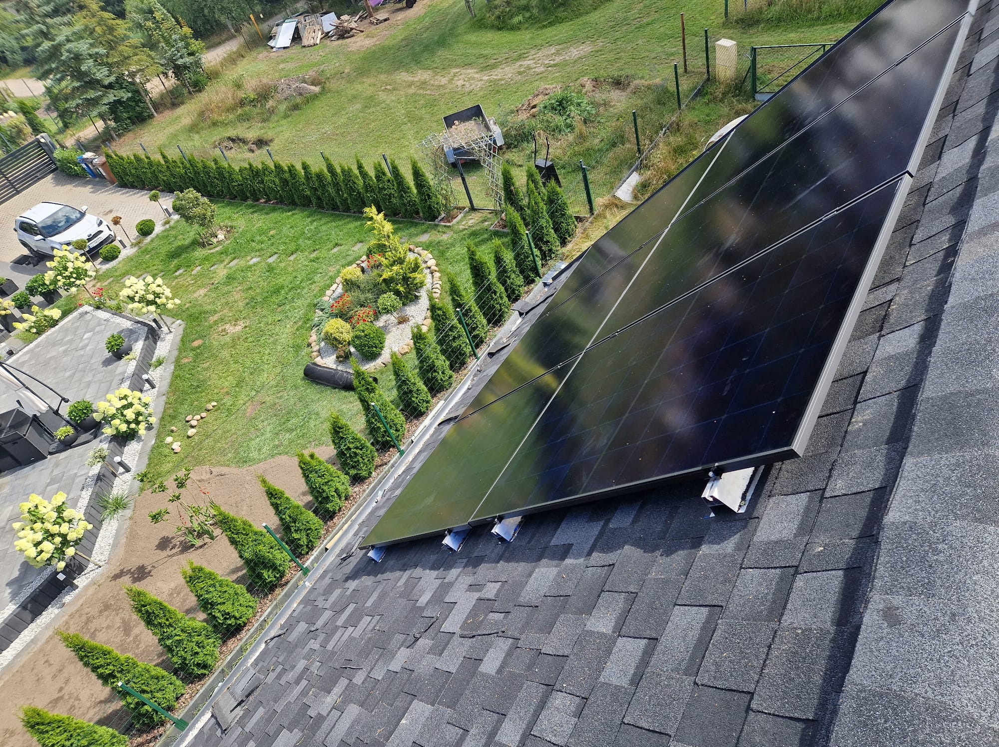 Instalacja paneli słonecznych na dachu domu z widokiem na zadbany ogród i podjazd. Dach pokryty gontem bitumicznym. Ujęcie z góry, szeroki kadr.