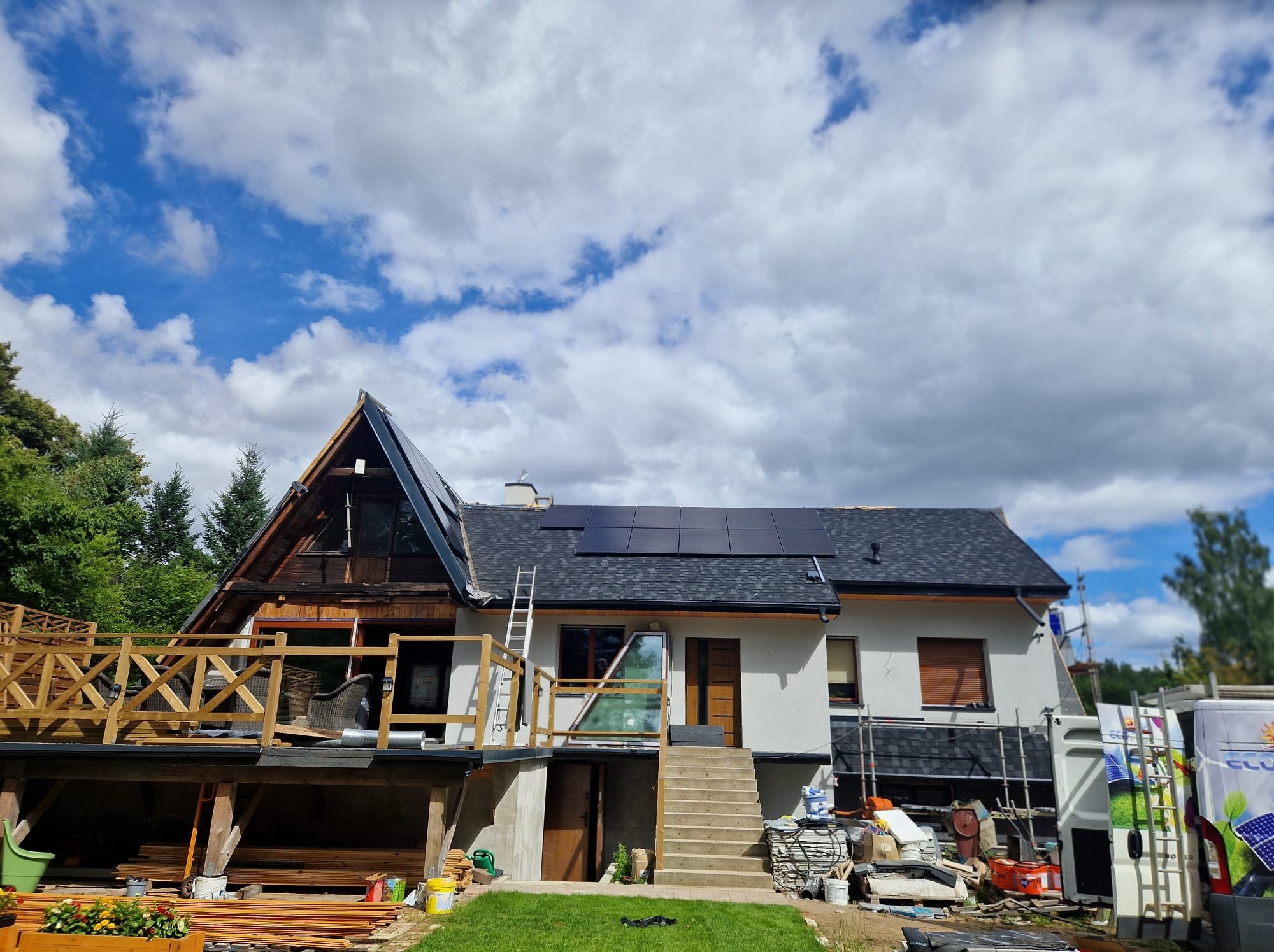 Dom z zainstalowanymi panelami słonecznymi na dachu, widoczne rusztowania i elementy montażowe. Częściowo ukończony montaż paneli na drugim dachu.