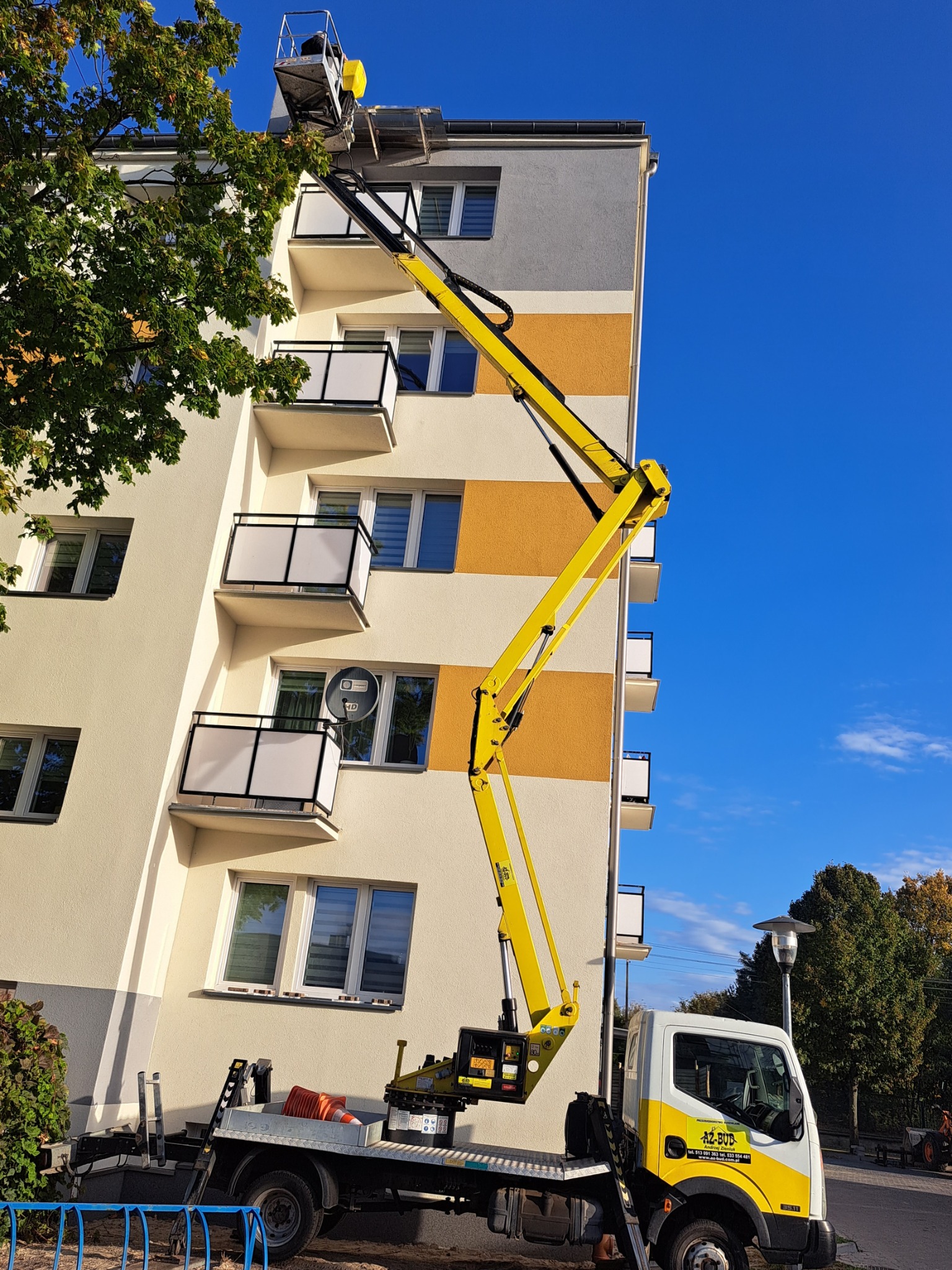 Pracownik na podnośniku koszowym przy elewacji bloku, widoczne balkony i odświeżona kolorystyka budynku w odcieniach beżu i pomarańczy.