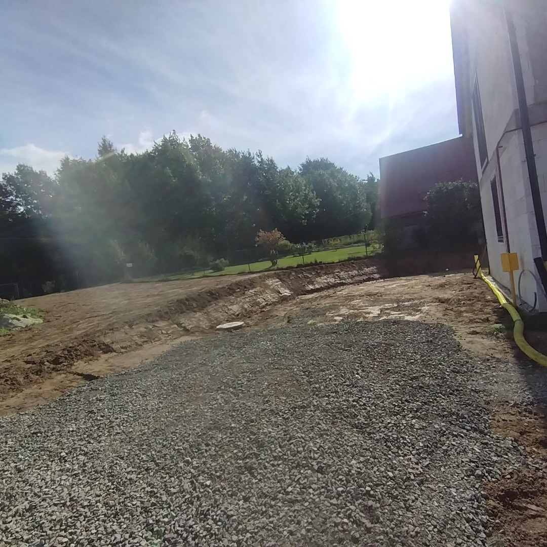 Teren po robotach ziemnych z widocznym wykopem i nasypem żwirowym, w tle zieleń drzew i budynek w stanie surowym.