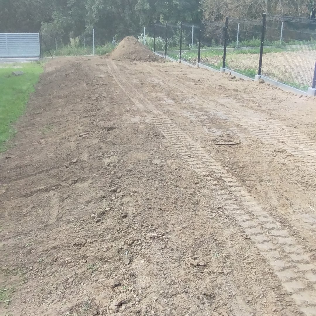 Przygotowany teren pod budowę drogi z widocznymi śladami opon maszyn budowlanych i stertą ziemi w tle, ogrodzenie po prawej stronie.