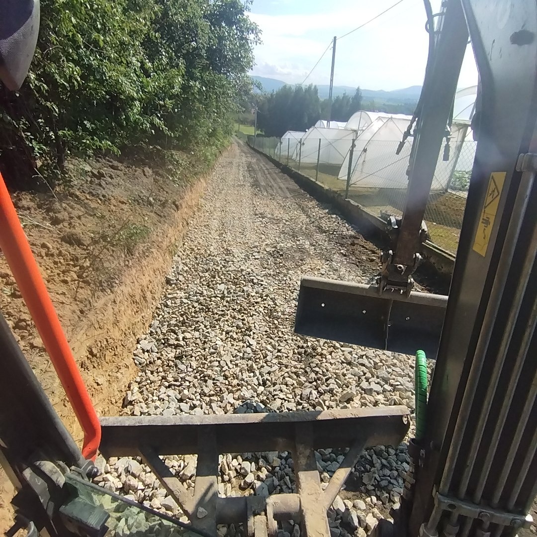 Widok z kabiny maszyny budowlanej na drogę z utwardzoną nawierzchnią żwirową, w tle szklarnie i zalesione wzgórza.