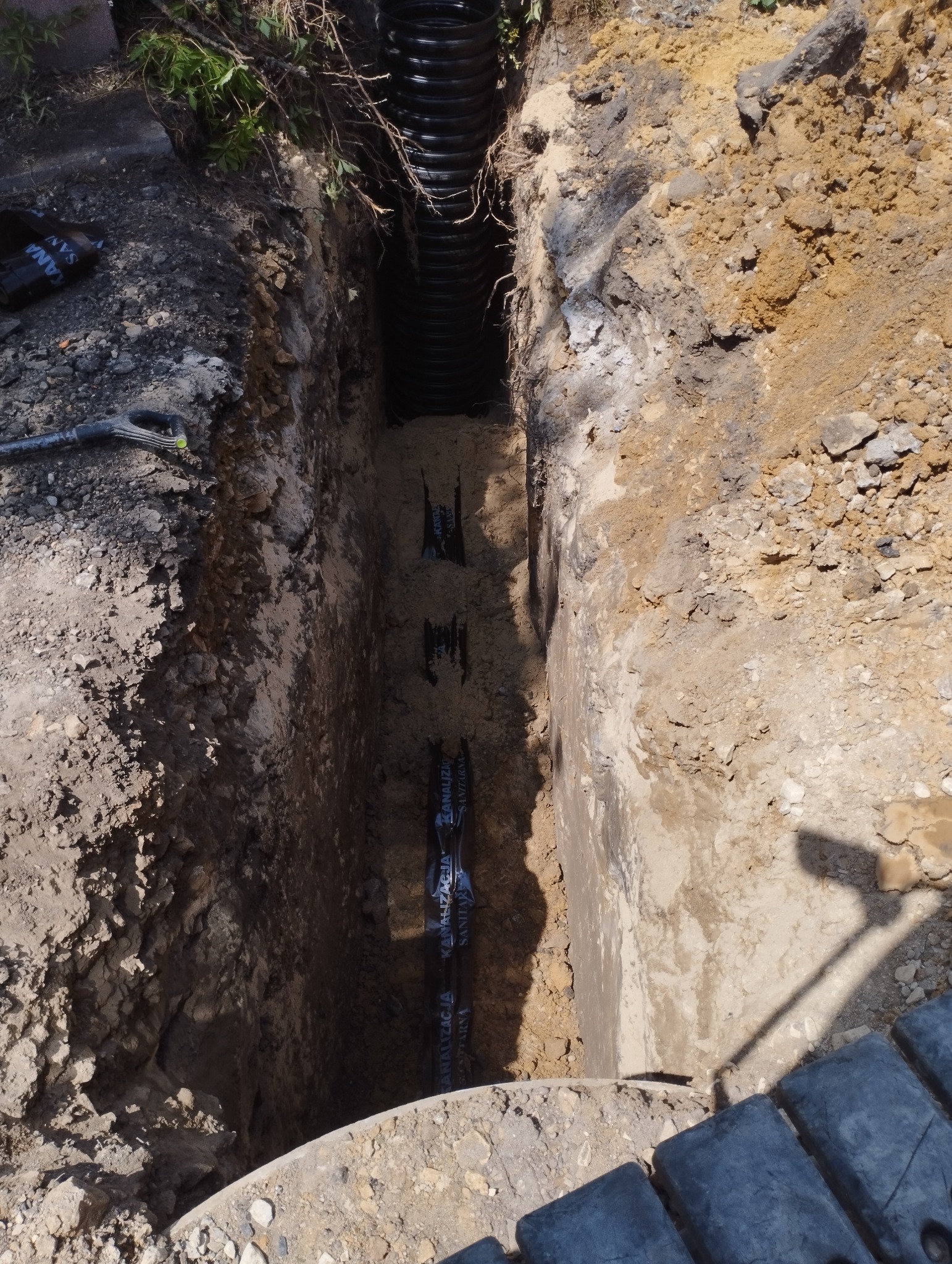 Wykop z ułożoną rurą kanalizacyjną z napisem 'kanalizacja sanitarna', widoczne ściany wykopu z różnymi warstwami ziemi i kamieni, fragment łopaty i gąsienic koparki w kadrze.