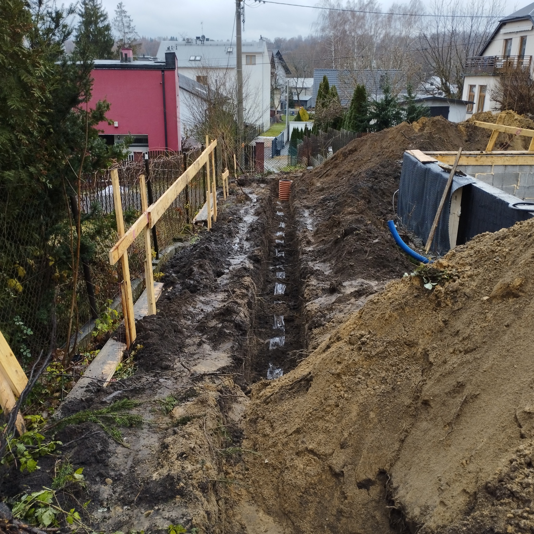Wykopany wąski rów w ziemi z widocznymi studzienkami kanalizacyjnymi, przygotowany do instalacji, z drewnianym ogrodzeniem w tle i zabudową mieszkalną.