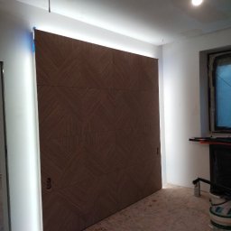 PRIMEBUD NAZAR KACHAK - Panel ścienny z geometrycznym wzorem w odcieniach brązu, podświetlony listwą LED, tworzący nowoczesny akcent w jasnym wnętrzu. Widoczne detale wykończenia.