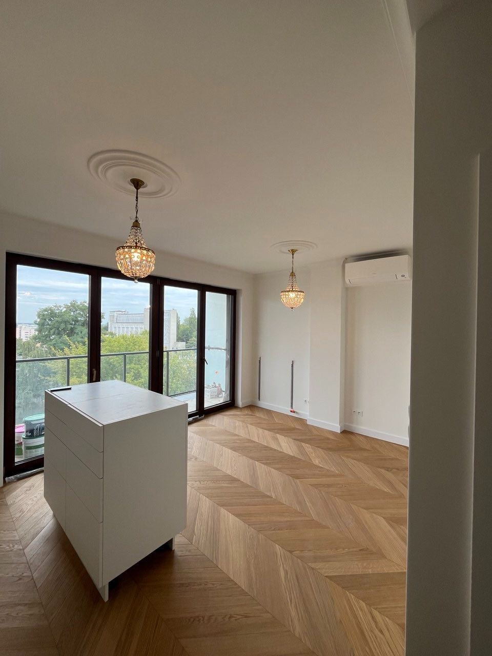 Jasne wnętrze z parkietem w jodełkę, białymi ścianami i dwoma kryształowymi żyrandolami. Widoczne duże okna z widokiem na zieleń i biała komoda. Minimalistyczny design.