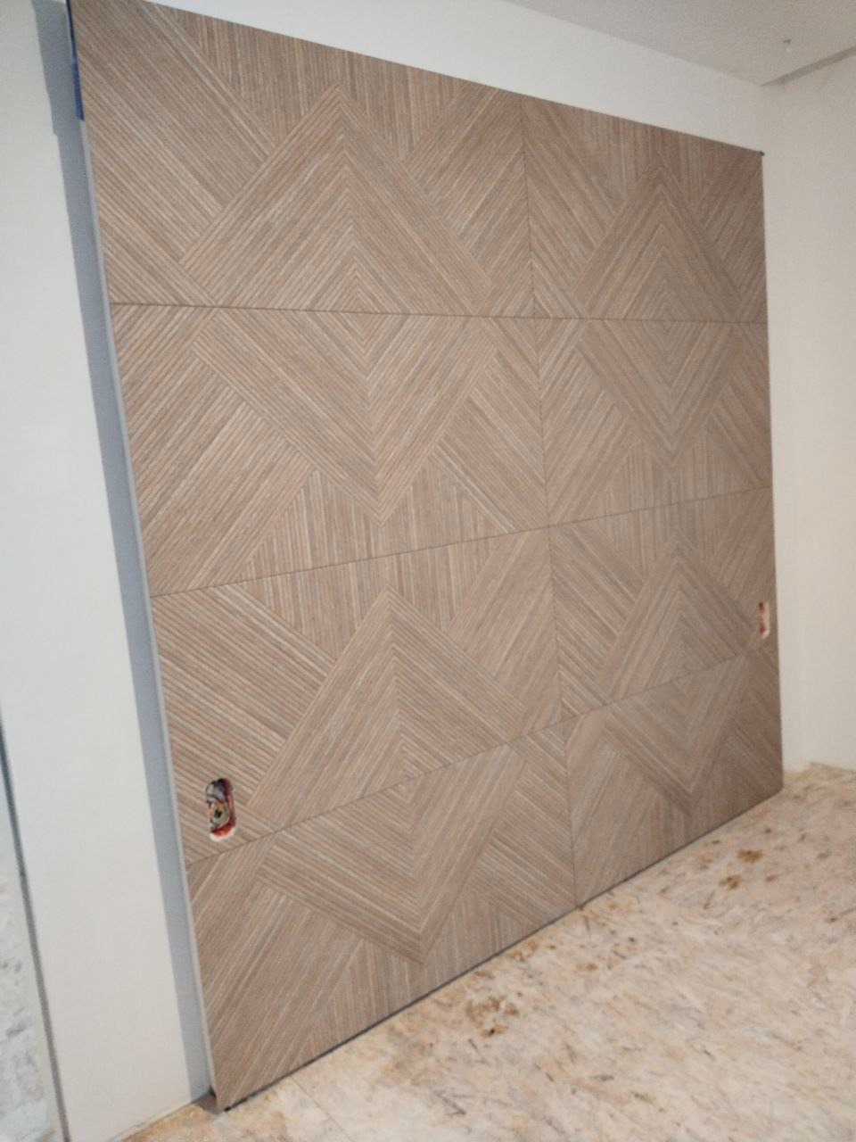 Ściana wyłożona płytkami z geometrycznym wzorem w odcieniach beżu i brązu. Widoczne gniazdka elektryczne. Ukośne ułożenie płytek tworzy unikalny efekt wizualny.